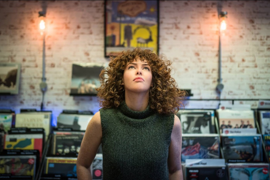 Woman with curly hair in a green sleeveless sweater stands among vinyl records in a record store, looking upward/artfully gazing at something above.