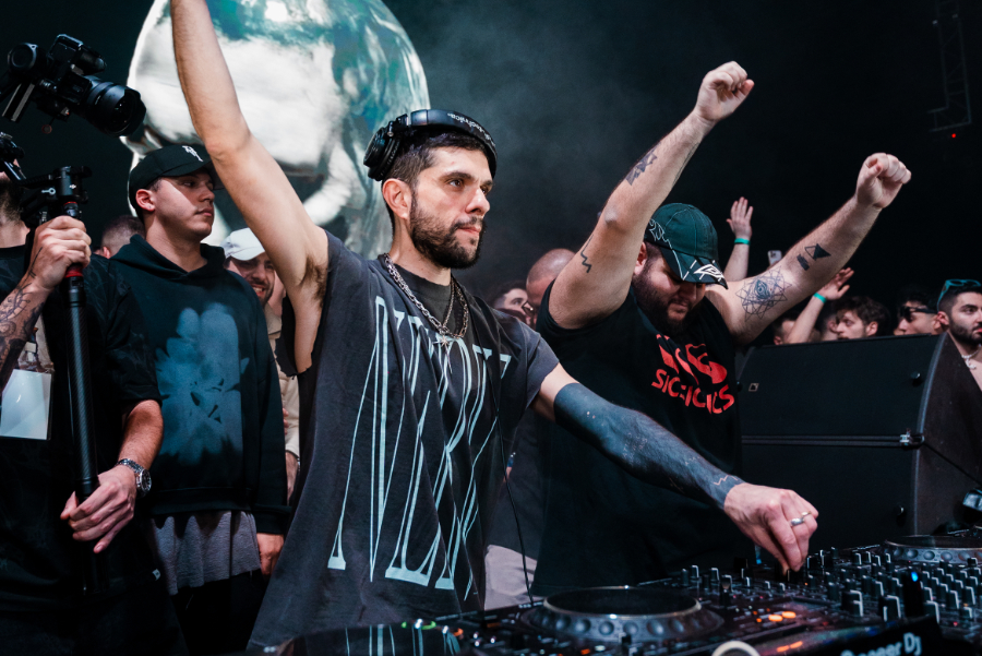 Two DJs perform at a club, one raising a fist while the crowd cheers behind them with cameras recording the moment, and a mixer in the foreground.