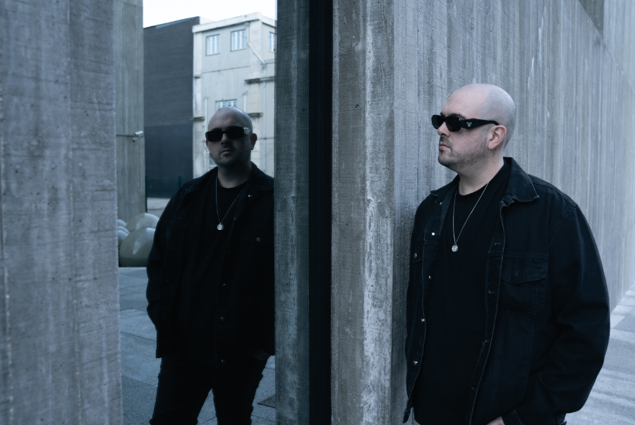 Bald man in a black jacket and sunglasses stands between concrete walls in an urban setting, with a reflection in glass.