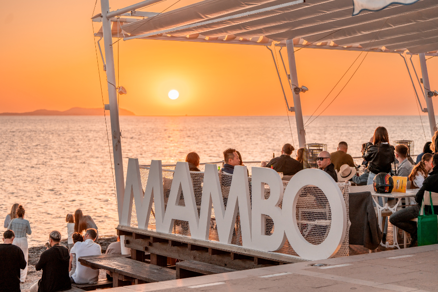 Sunset over the sea at a lively outdoor venue with a large white 'MAMBO' sign and groups of people seated at tables by the water.