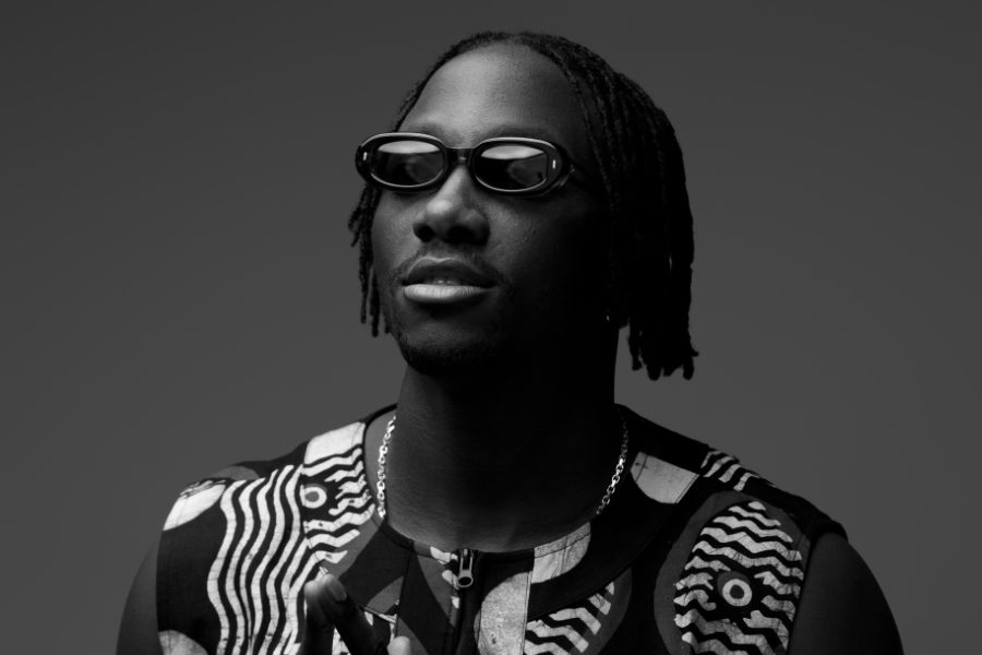 Portrait of a Black man wearing dark round sunglasses, a patterned sleeveless top, and a silver chain against a gray backdrop.