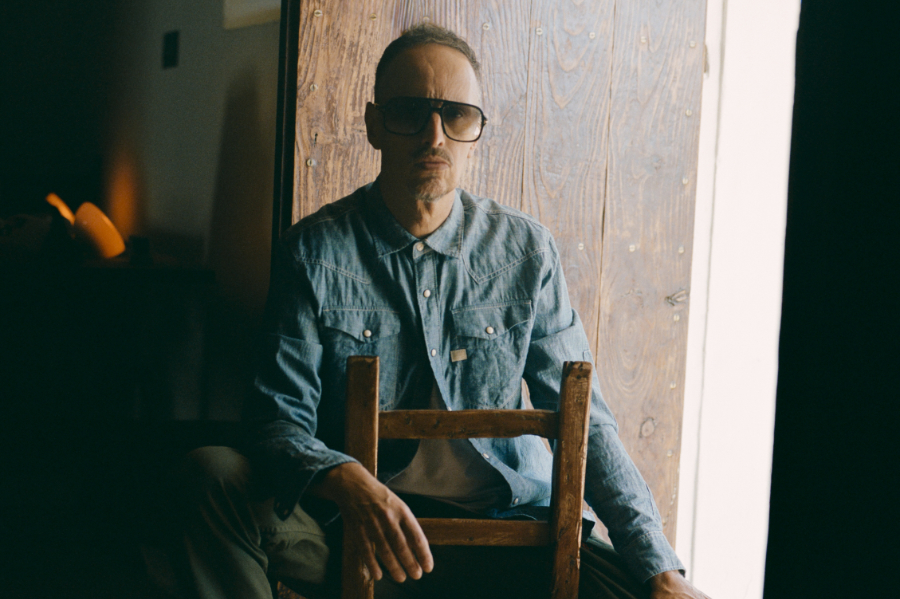 Man wearing sunglasses and a denim shirt sits in a doorway, hands resting on a wooden chair in front of him.