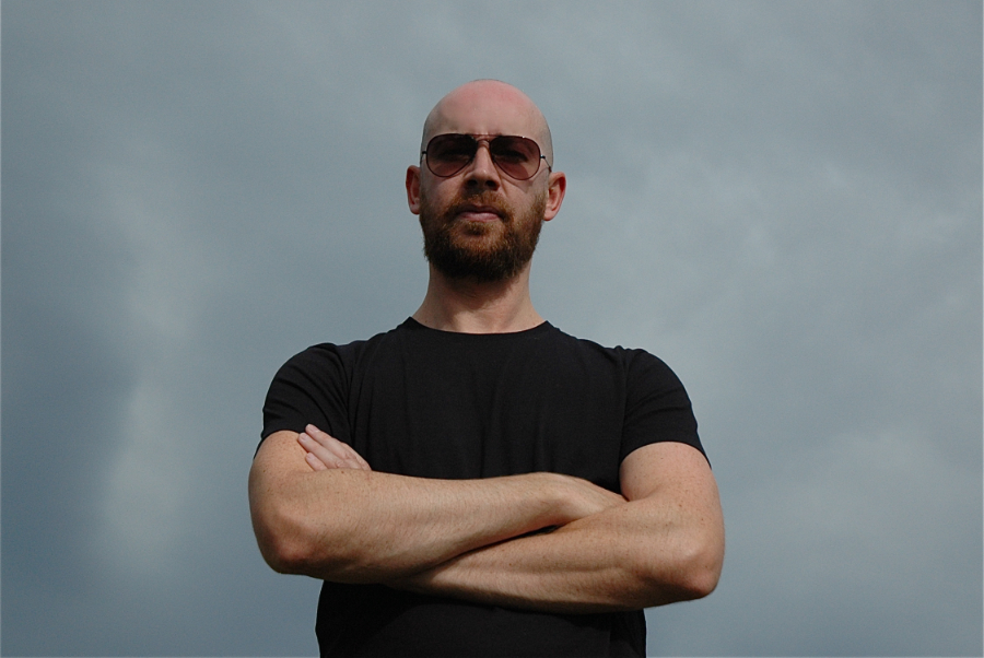 Bald man with beard wearing sunglasses and a black T‑shirt stands with arms crossed against a cloudy sky.