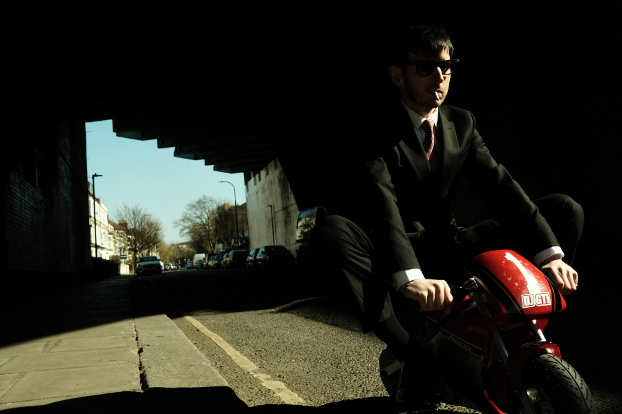 Man in a suit and sunglasses sits on a small red motorcycle in a dark underpass, smoking a cigarette.