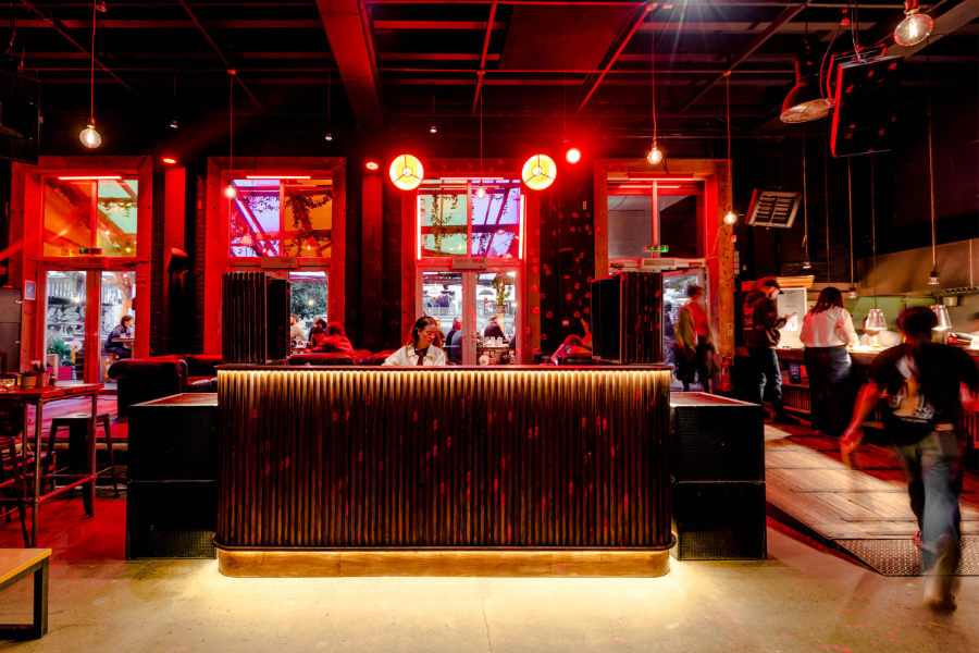 Bar interior at night with red lighting, a long ribbed wooden counter in the center, and patrons seated around it.