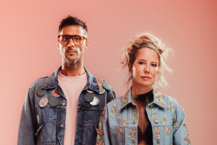 Man and woman stand against a pink backdrop wearing patched denim jackets, facing the camera.