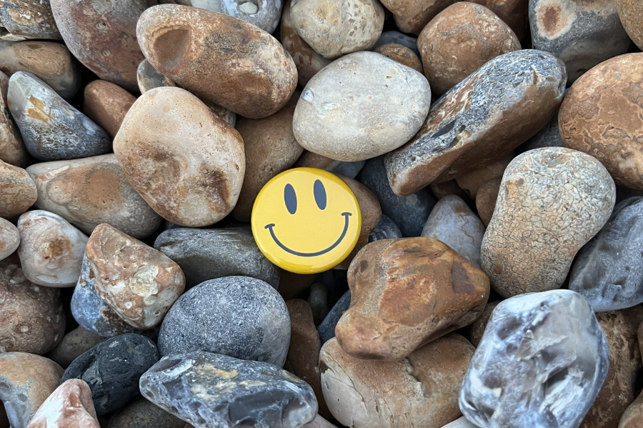Yellow smiley-face button resting on smooth brown and gray rocks with pebbles nearby.