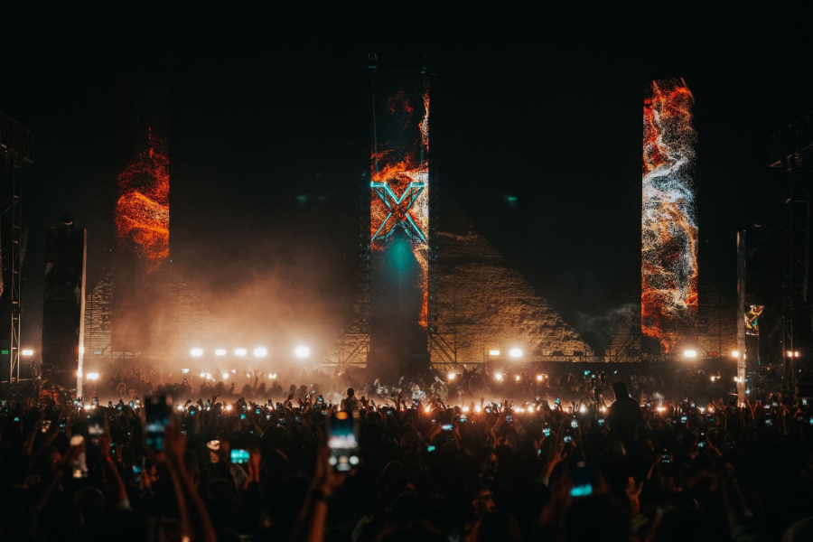 Nighttime outdoor concert with a packed crowd, bright stage lights, and tall vertical video pillars displaying fiery visuals.