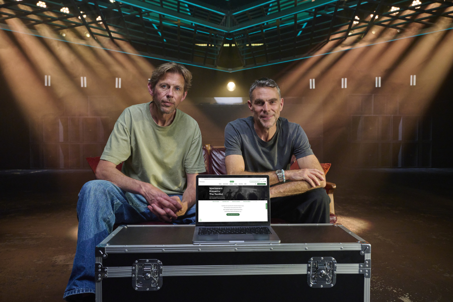 Two men sit on a couch behind a laptop on a gear case in a dim studio with stage lights overhead, looking at the camera.