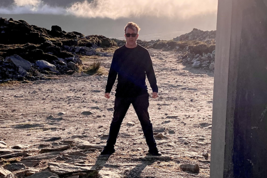 Man in black clothing and sunglasses stands with legs apart on a rocky beach at sunset.