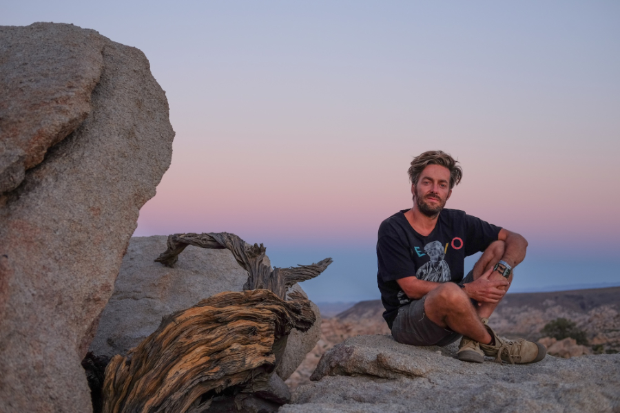 Man sitting cross-legged on a large rock in a desert at sunset, with rugged terrain and a weathered log nearby.
