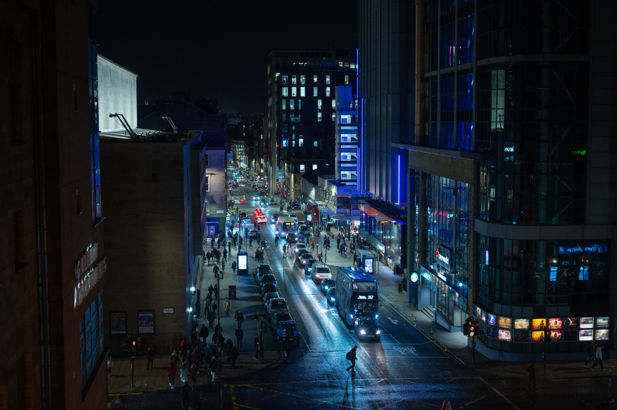 Nighttime city street filled with cars, buses, and pedestrians along a lit modern downtown avenue. (informative)