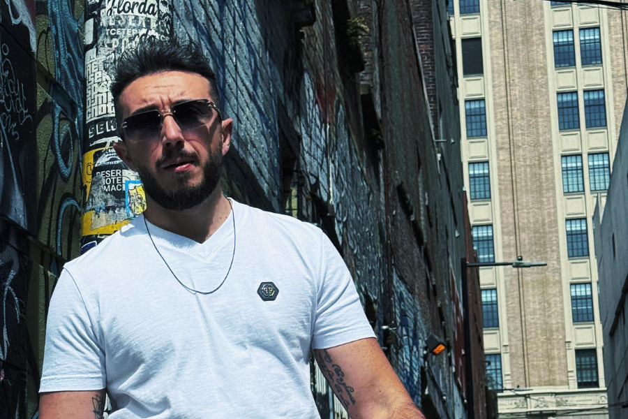 Man with dark hair and beard wearing a white t-shirt and sunglasses stands in a graffiti-covered alley.