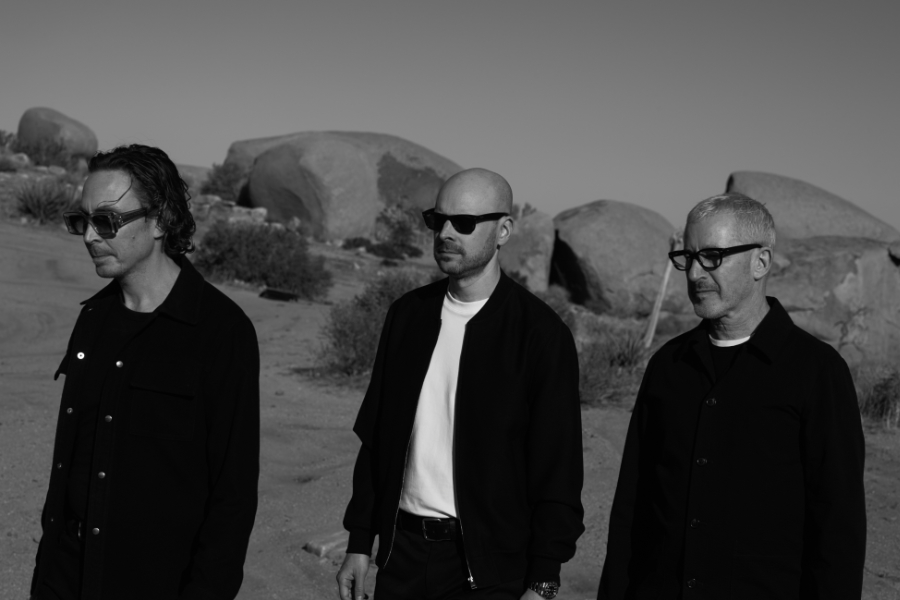 Three men in black jackets and sunglasses stand in a rocky desert landscape, posing for a photo in grayscale.