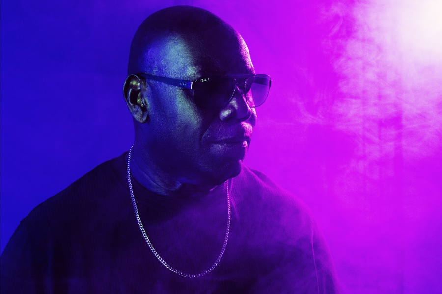 Man wearing dark sunglasses and a silver chain necklace, lit by purple and blue stage lights with smoke around him.